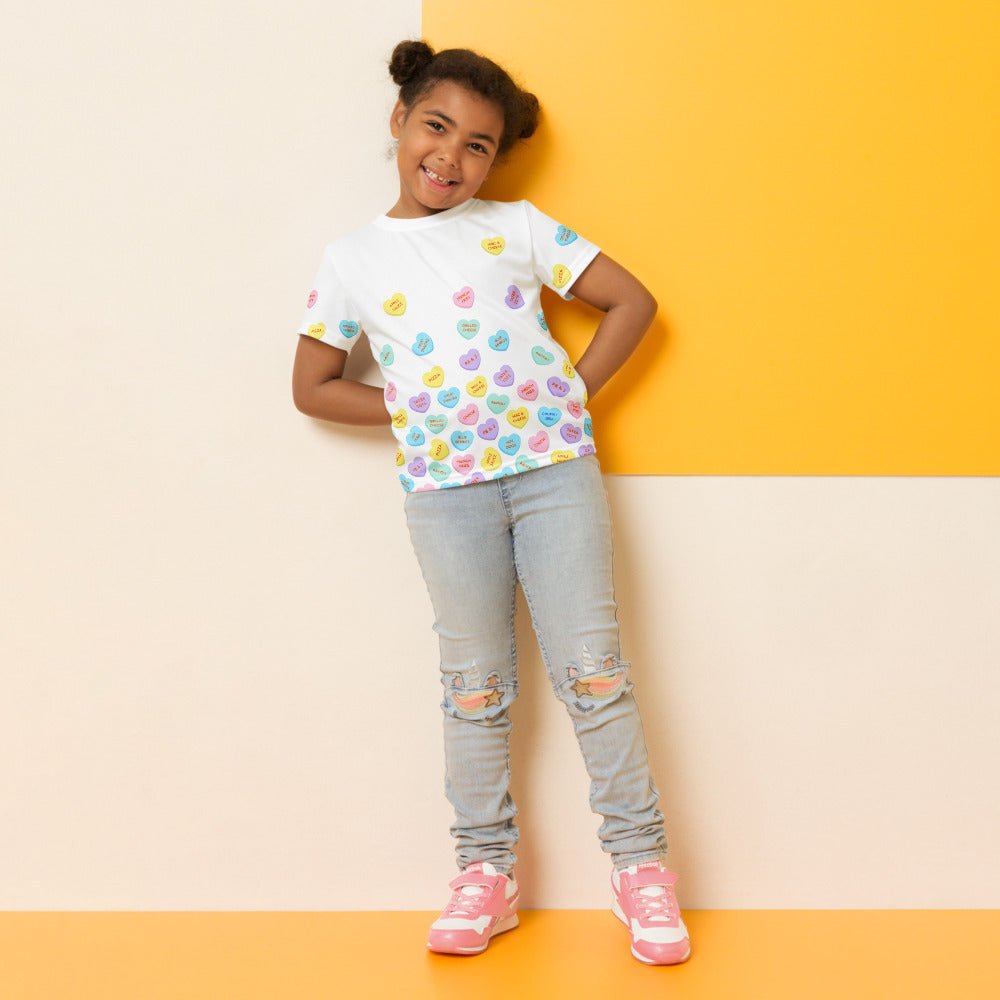 Chunky Deli product image: Smiling girl wearing the "Candy Hearts" t-shirt (white with colorful candy hearts featuring food illustrations), jeans, and tennis shoes, standing in front of a yellow wall.