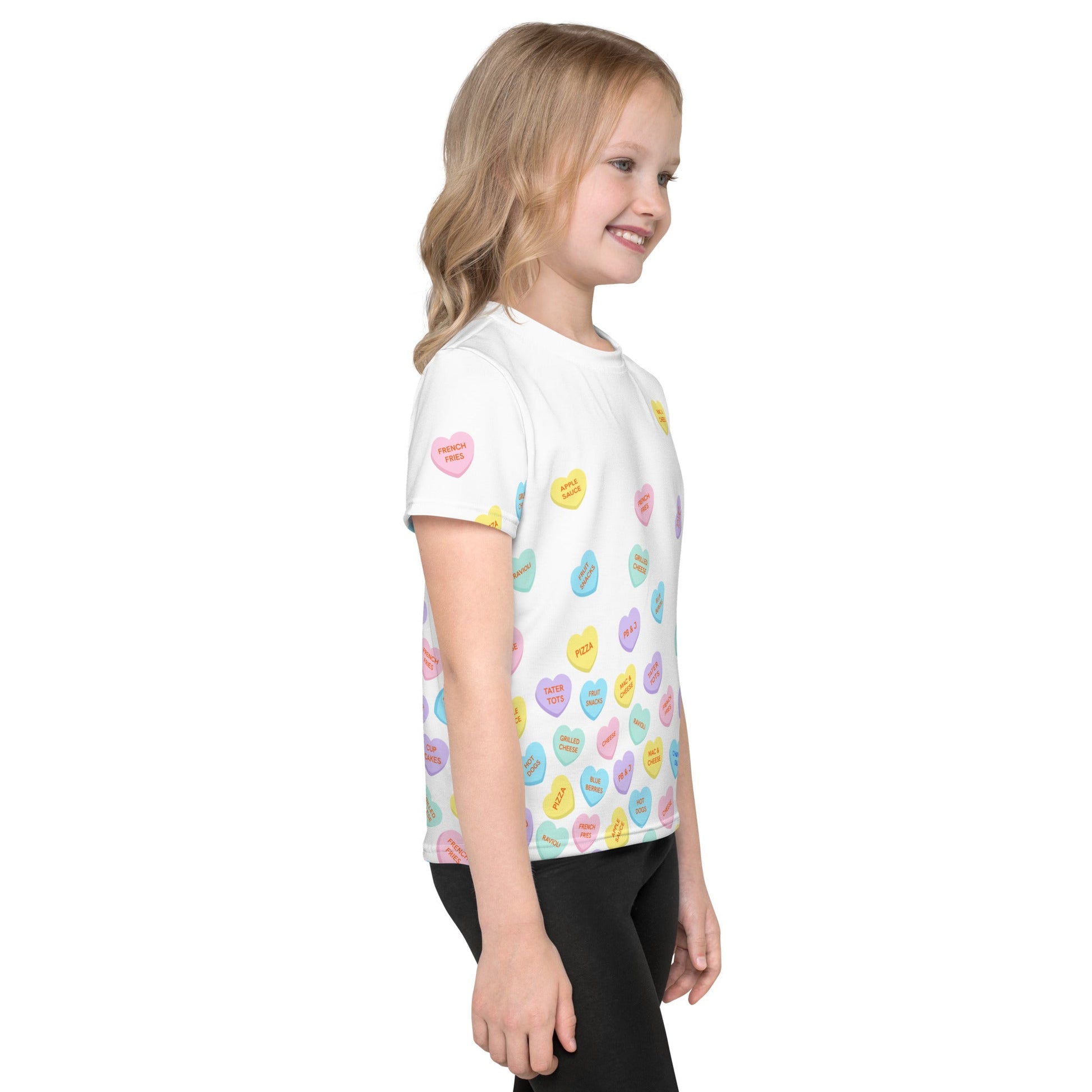 Chunky Deli product image: Right side view of a girl wearing the "Candy Hearts" t-shirt (white with colorful candy hearts featuring food illustrations), showing the design on the side of the shirt. She is wearing black pants and standing against a white b