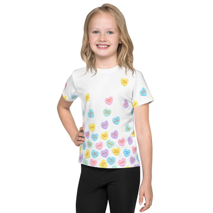 Chunky Deli product image: Close-up of a smiling girl wearing the "Candy Hearts" t-shirt (white with colorful candy hearts featuring food illustrations), with one hand on her hip and wearing black pants, against a white background.