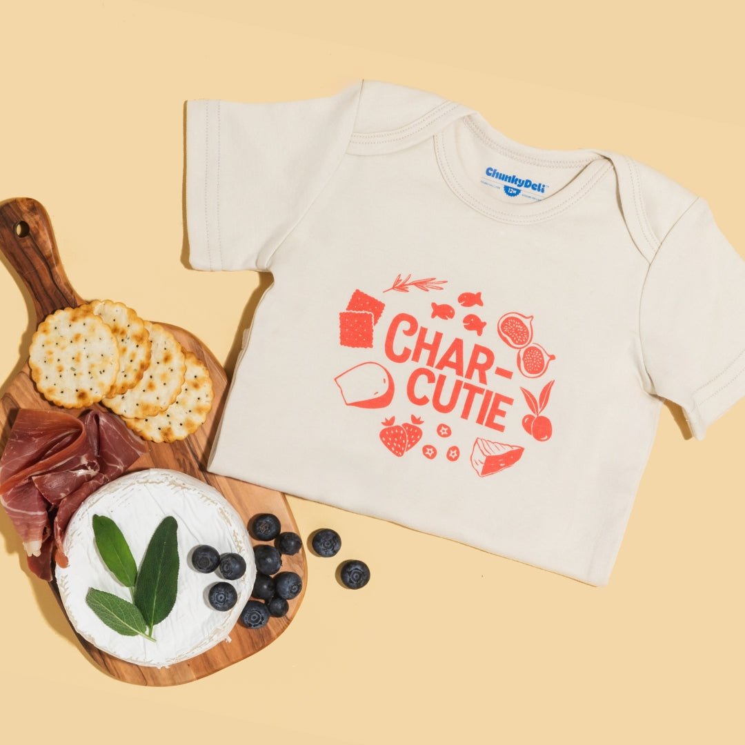 Chunky Deli product image: "Charcutie" onesie (100% cotton, natural color) folded in half next to a charcuterie board featuring olives and bread, playing off the onesie's graphic design. Peach background.