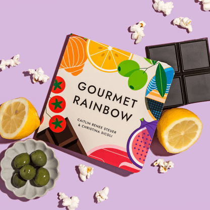 "Gourmet Rainbow" board book displayed at an angle among gourmet foods such as olives, oranges, chocolate, and popcorn on a purple background. Chunky Deli product image.