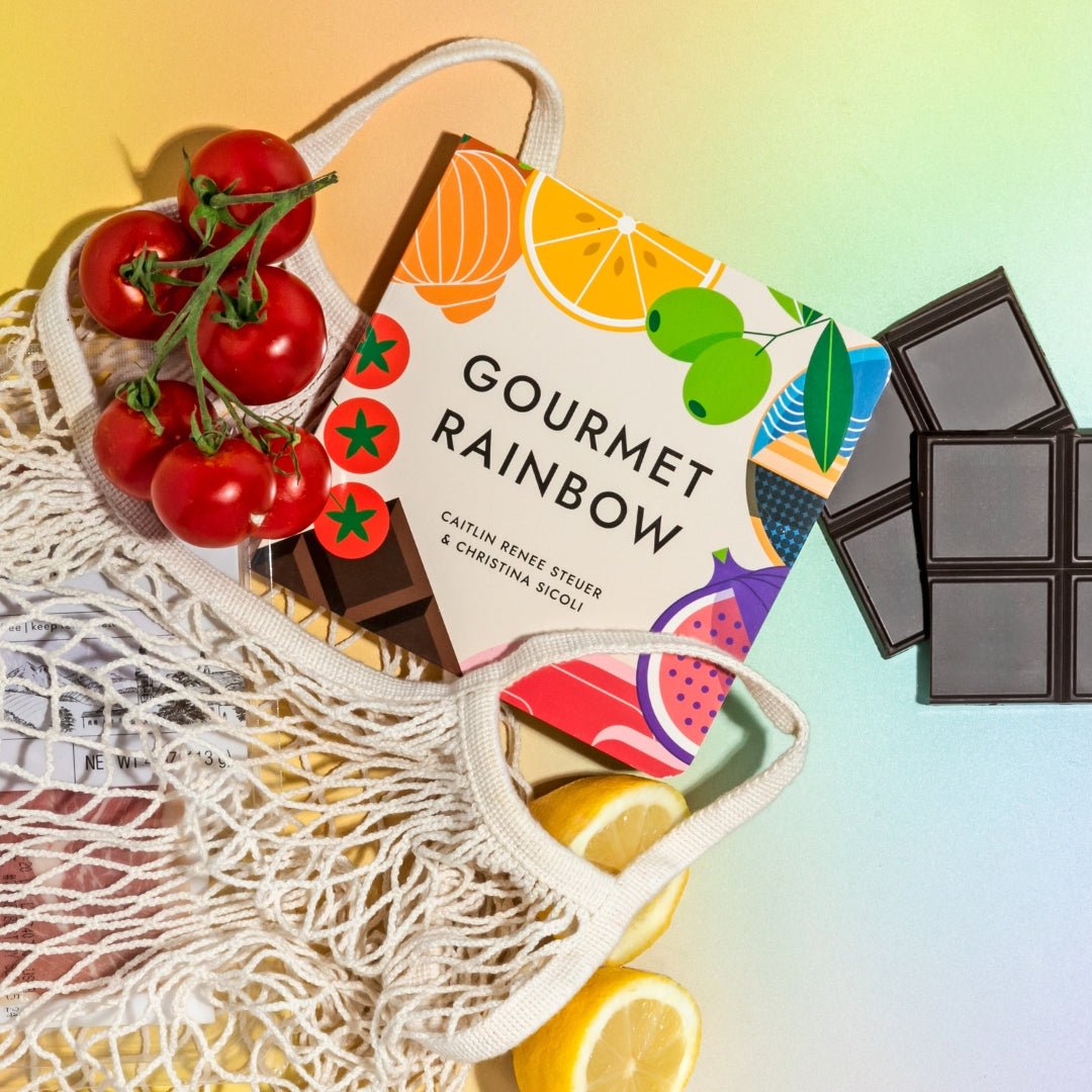 "Gourmet Rainbow" board book popping out of a grocery bag, surrounded by food items like tomatoes and chocolate on a rainbow gradient background. Chunky Deli product photo.