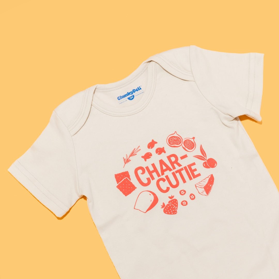  Close-up of the "Charcutie" baby onesie in natural cotton on an orange background. The graphic features the word "Charcutie" surrounded by food illustrations. Chunky Deli product image.