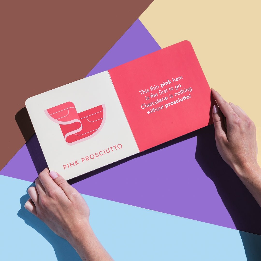 Two hands holding the "Gourmet Rainbow" board book open to a page featuring pink prosciutto on a colorful background. Chunky Deli product image.