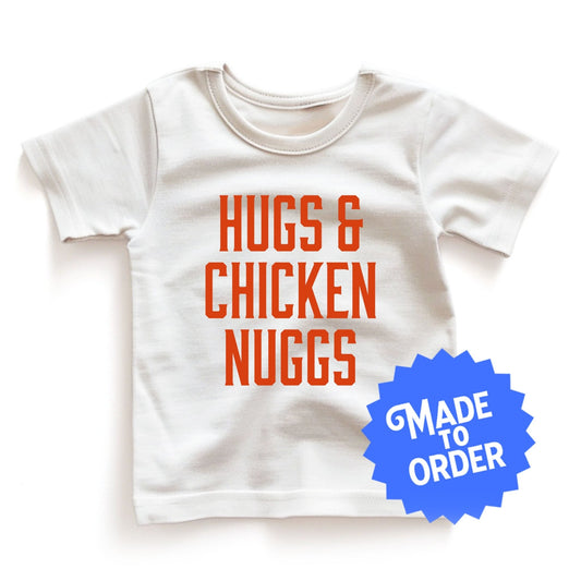 Chunky Deli product image: White short-sleeved kids' and toddler t-shirt with the text "Hugs and Chicken Nugs" stacked vertically in orange font on the chest.