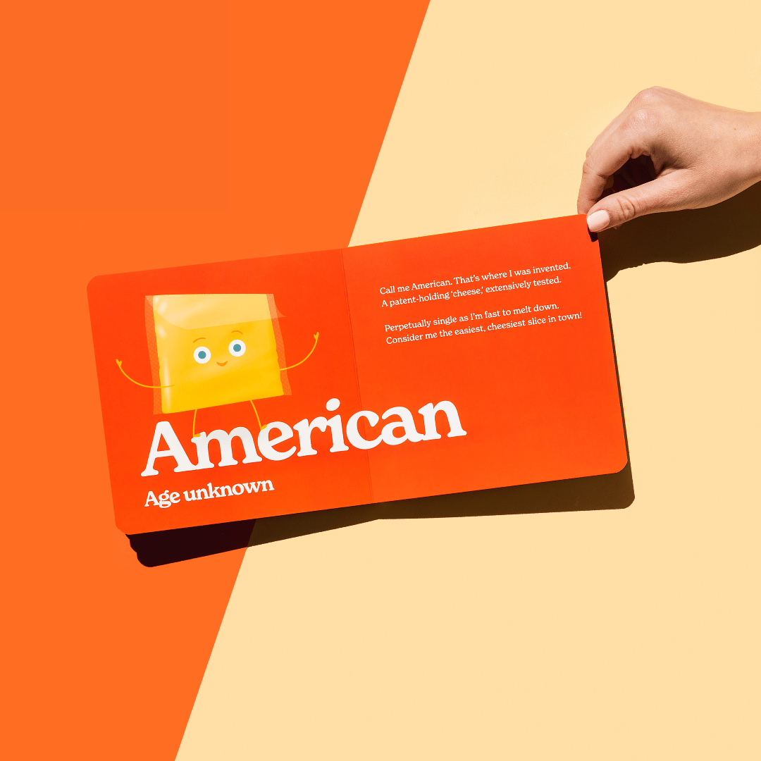 Chunky Deli product image: "Little Book of Cheese" open to the American cheese page, featuring an illustration of a cheese slice and its accompanying playful rhyme, held open by one hand against an orange background.