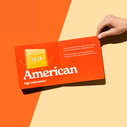 Chunky Deli product image: "Little Book of Cheese" open to the American cheese page, featuring an illustration of a cheese slice and its accompanying playful rhyme, held open by one hand against an orange background.