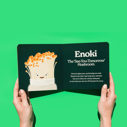 Chunky Deli product image: "Little Book of Fungi" open to the Enoki mushroom page (dark green spread), featuring an illustration of the Enoki mushroom character, held by two hands against a bright green background.