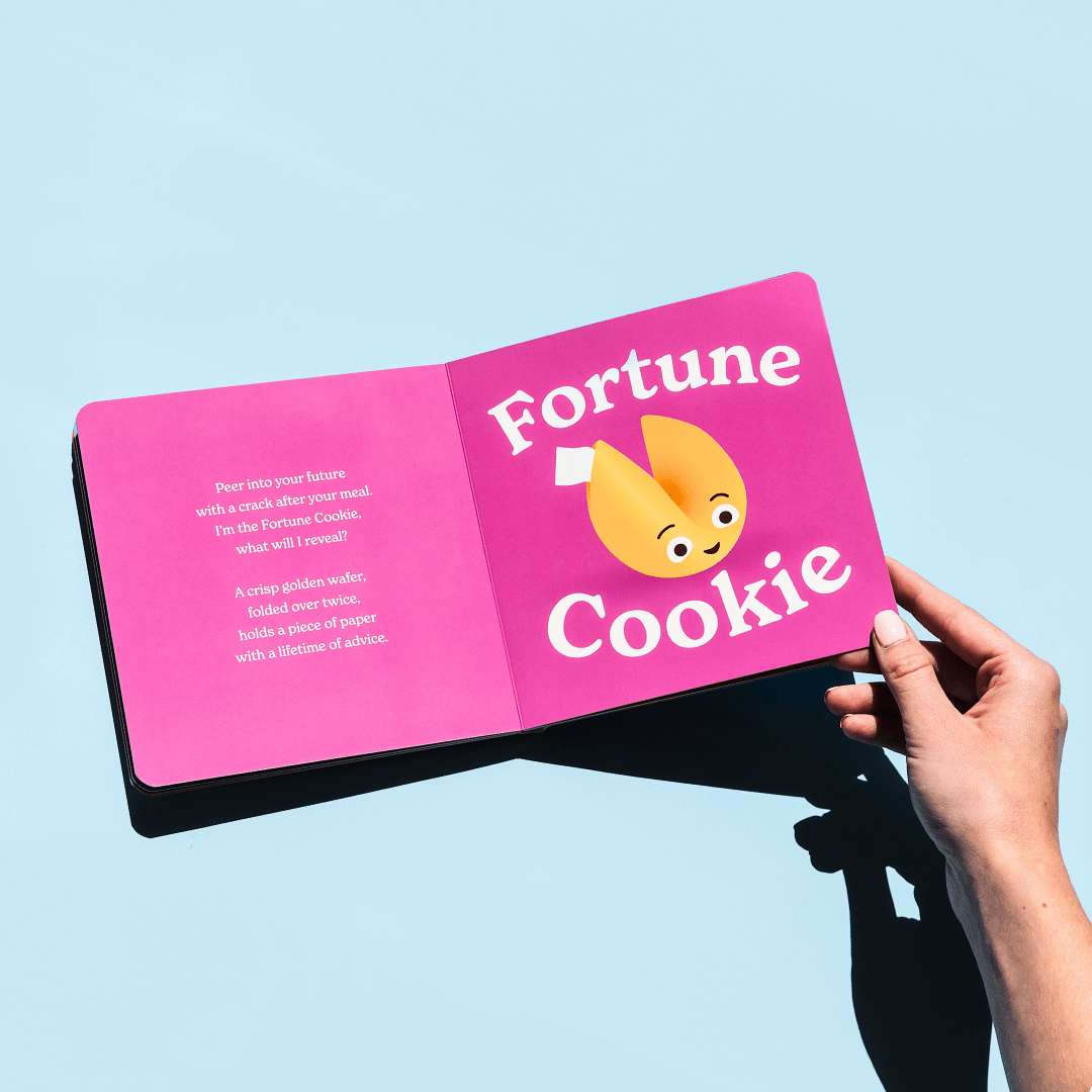 Chunky Deli product image: "Little Book of Cookies" open to the fortune cookie spread (purple page) featuring an illustration of a fortune cookie and accompanying rhyme, held open by one hand against a light blue background.