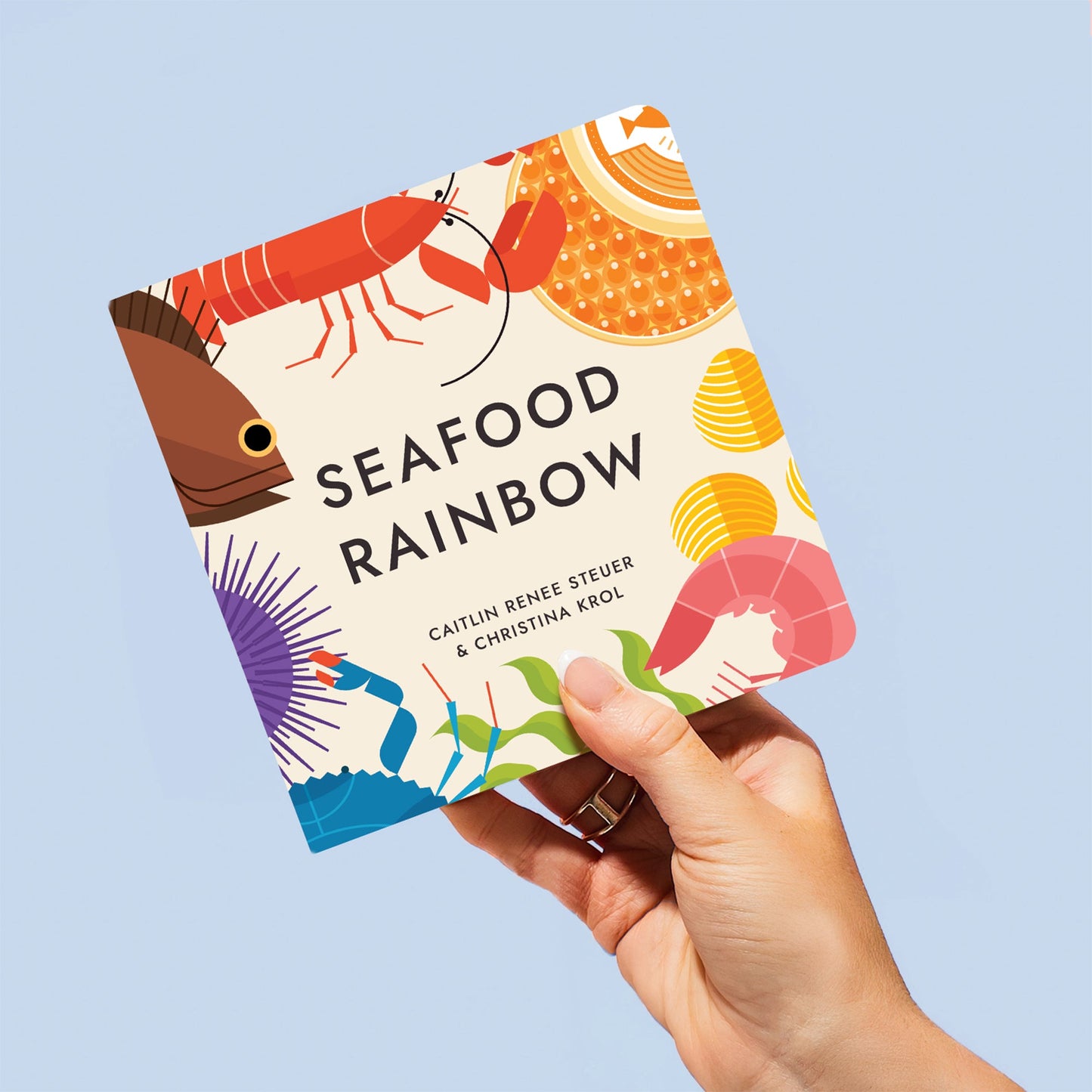 "Seafood Rainbow" book cover featuring vibrant seafood illustrations, held in a hand against a light blue background. Chunky Deli product image.