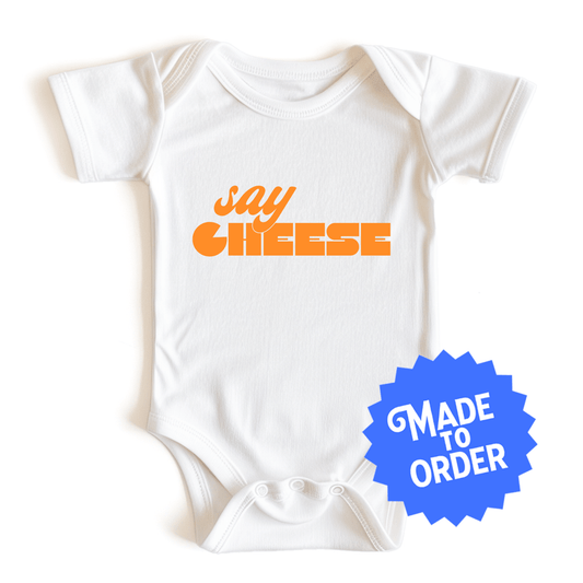 Chunky Deli product image: Natural-colored "Say Cheese" icon onesie, 100% cotton with envelope shoulders and three-snap closure, laid flat on a white background.