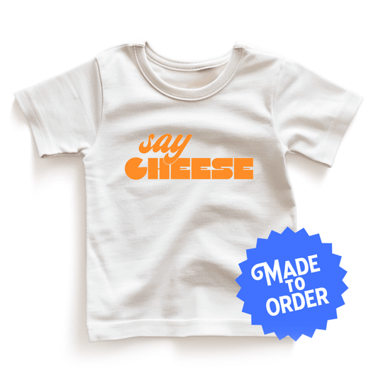 Chunky Deli product image: White short-sleeved kids' and toddler t-shirt with the text "Say Cheese" in cursive font across the chest, laid flat on a white background.