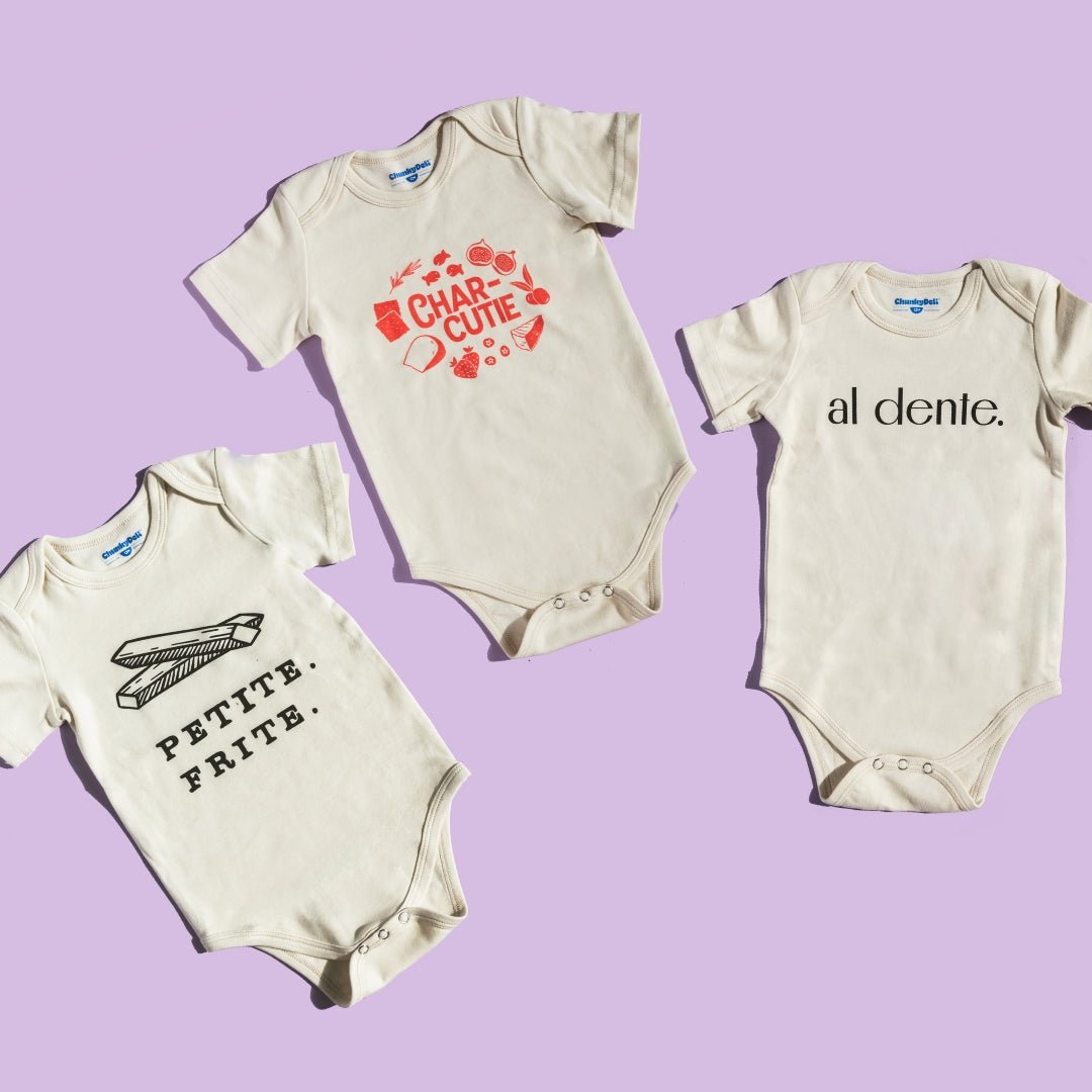 Chunky Deli product image: Three natural-colored onesies fanned out on a purple background, each displaying a different graphic design: "Petite Frite," "Charcutie," and "Al Dente."