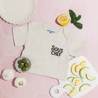 Chunky Deli product image: "Sous Chef" onesie folded in half and laid on a pink background, surrounded by real food items in a playful arrangement.