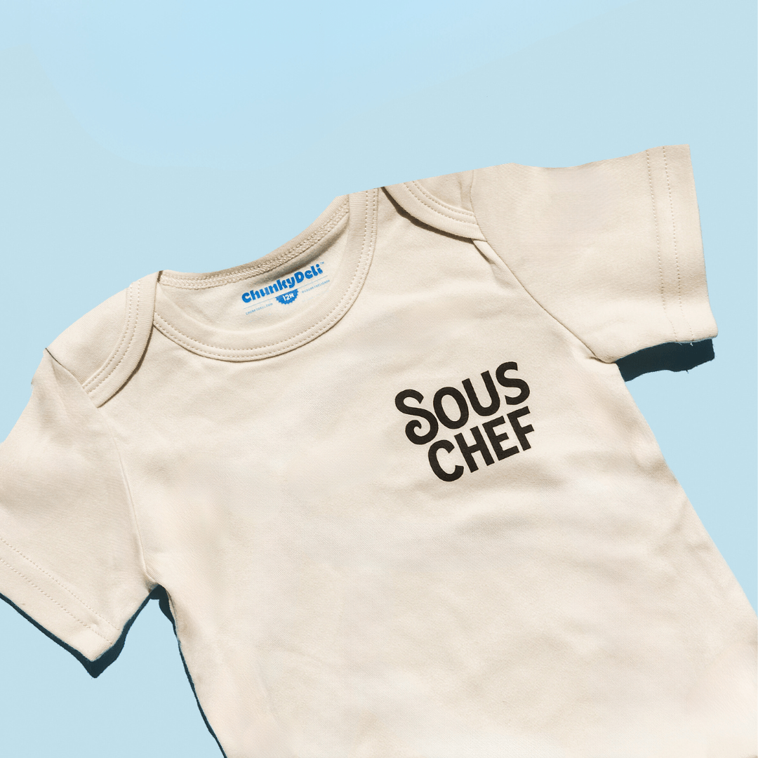 Chunky Deli product image: Close-up of the upper half of the "Sous Chef" baby bodysuit laid flat against a blue background, showcasing the design details.